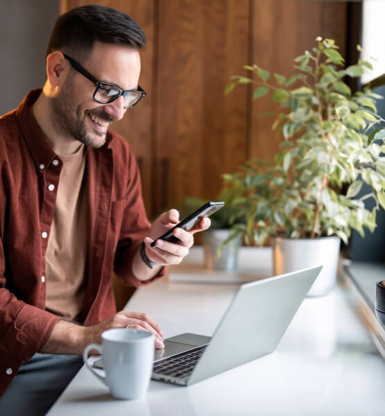 Man smiling while working on his laptop while holding his smart phone